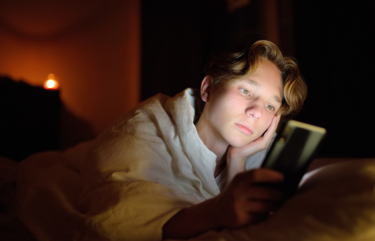 Teenage boy laying in bed on iphone resting his head on his hand