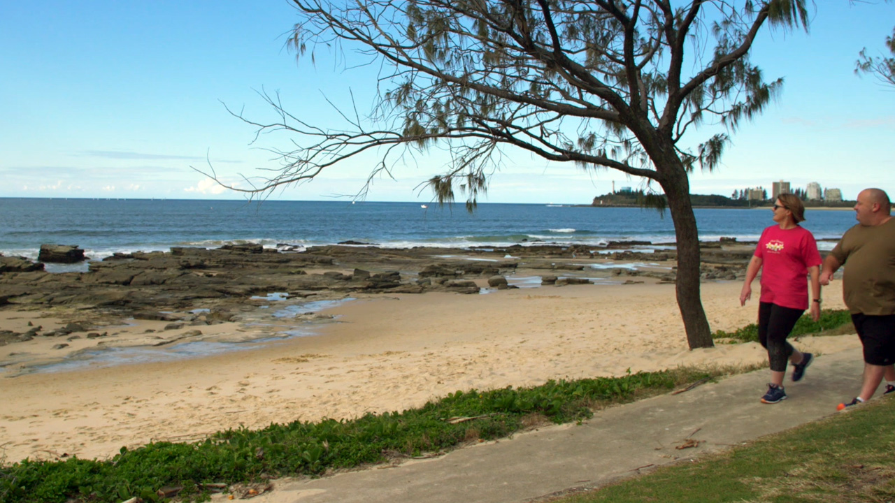 An empty beach with the tide out and a man in a green shirt and women in a pink shirt walking along the path looking out to the view